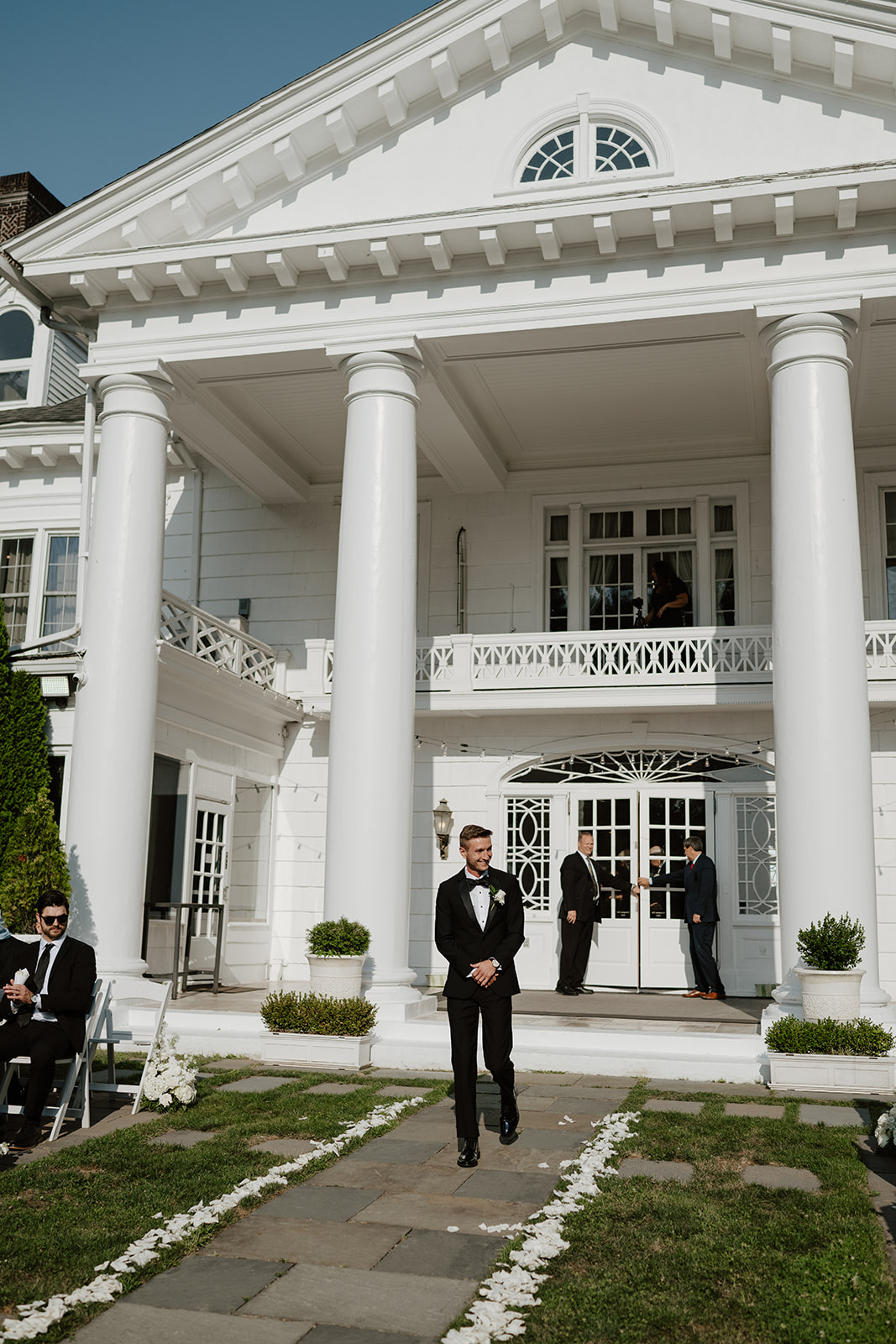 Groom smiles while walking down the flower-lined aisle in front of a grand white mansion with columns.